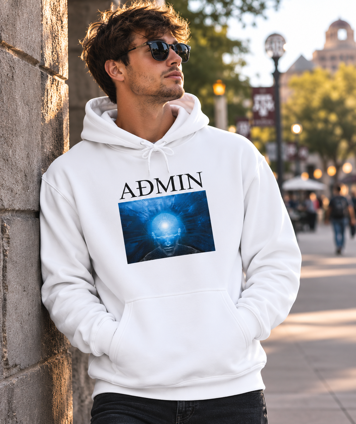 A man wearing a white pullover hoodie with a graphic print and the word 'ADMIN' stands against a stone wall in an outdoor urban setting.