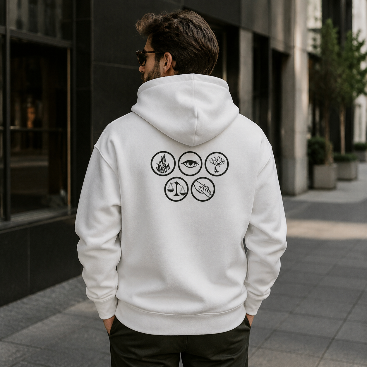 A man viewed from behind wearing a white pullover hoodie with a black graphic print of six circular symbols on the back, standing on a city sidewalk.