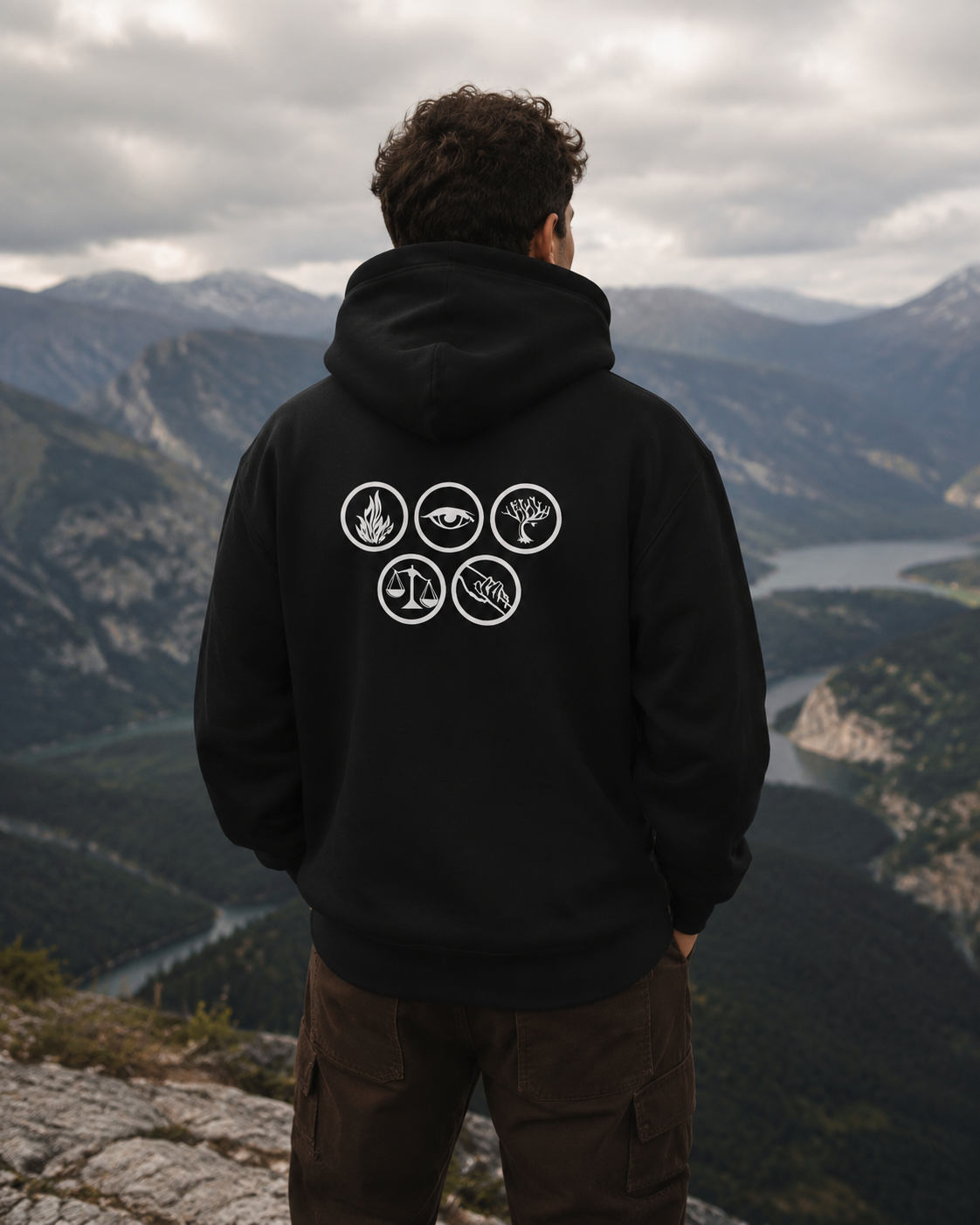 A man viewed from behind wearing a black hoodie with five white circular icons printed on the back, standing on a rocky mountain overlook with a vast valley and river landscape in the background.