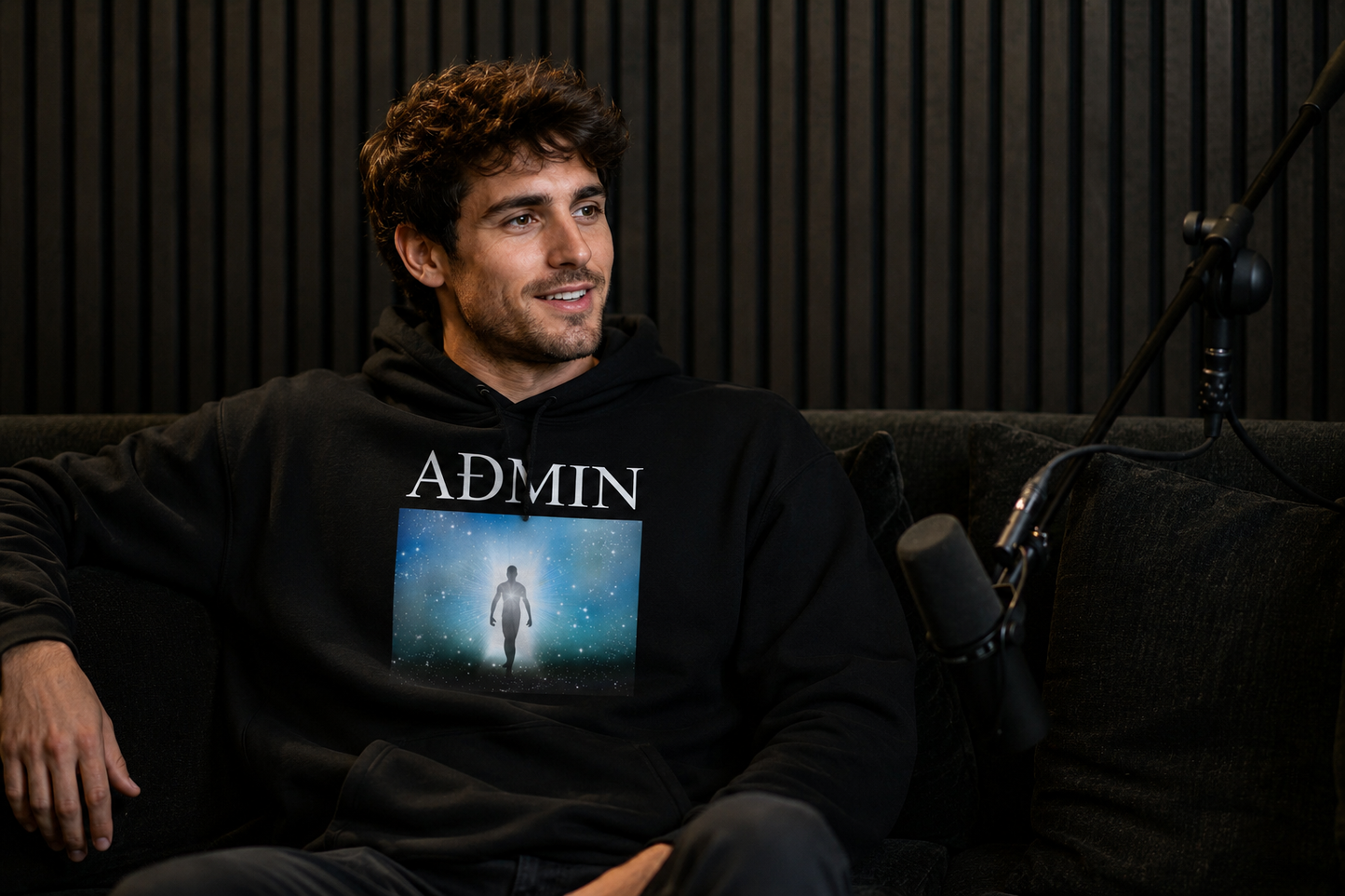 A man sitting on a dark couch wearing a black hoodie with the word 'ADMIN' and a graphic print on the front, set against a dark wood-paneled background with a microphone visible.