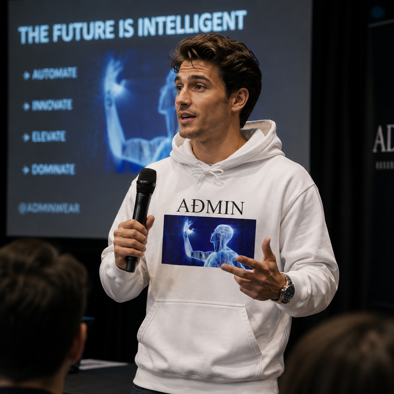 A man wearing a white hoodie with a graphic print and the word 'ADMIN' stands on a stage holding a microphone in front of a presentation screen.