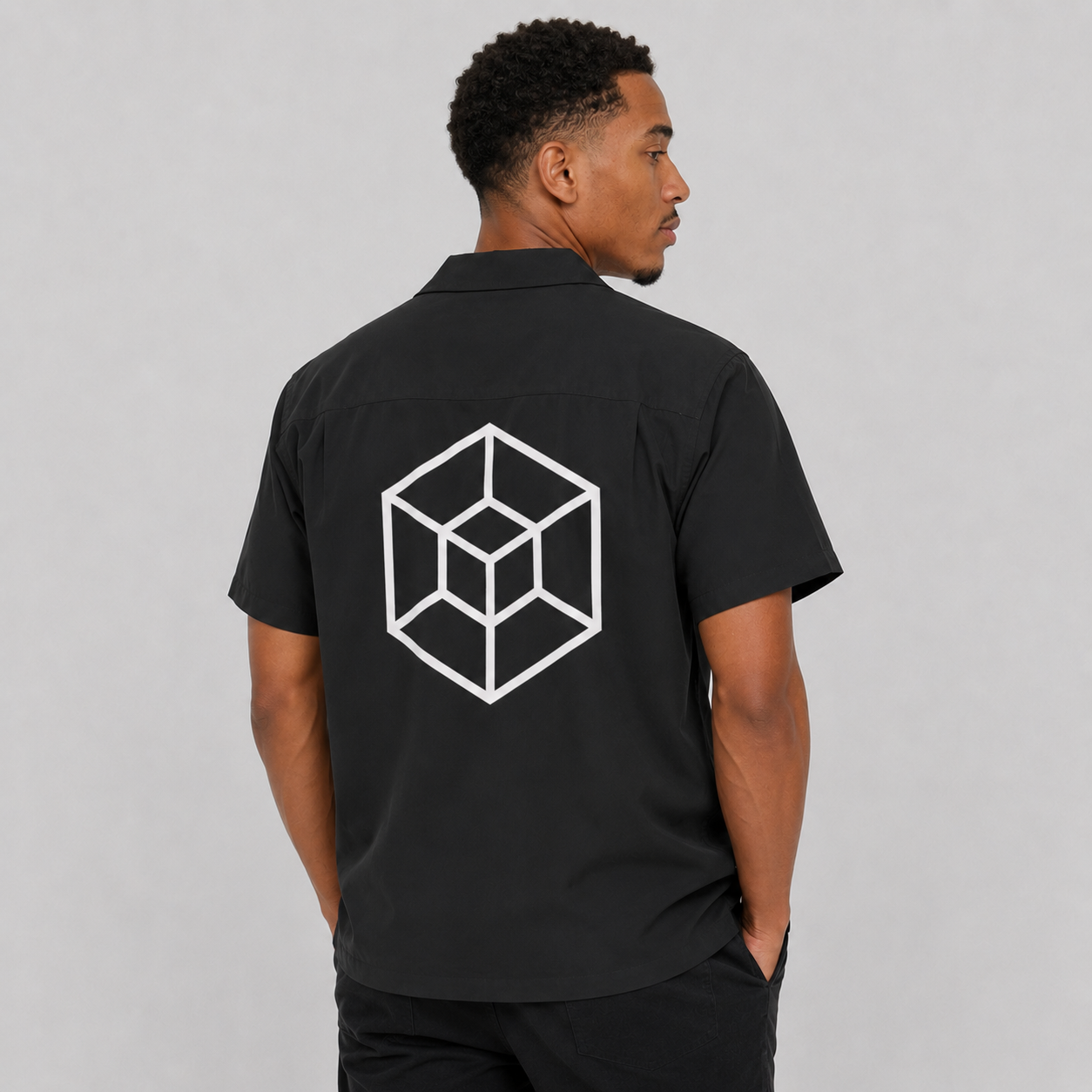 A man wearing a black short-sleeve button-up shirt, viewed from the back. The shirt features a large white geometric cube logo printed on the back.