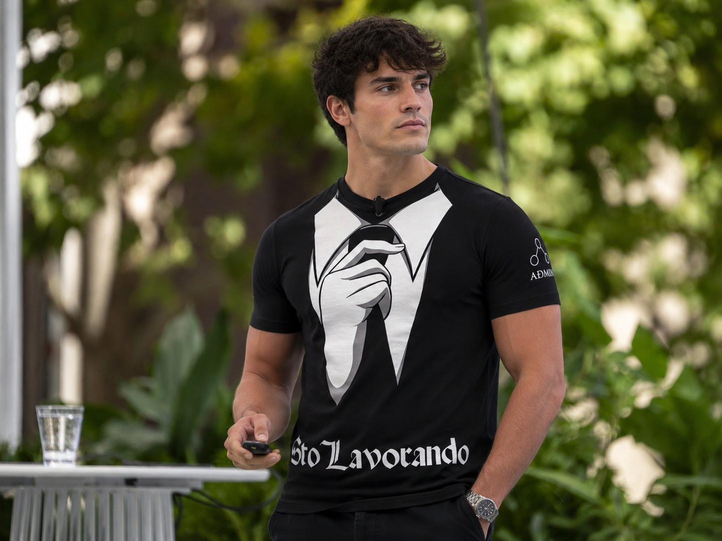 A man wearing a black t-shirt with a white graphic of a hand adjusting a bow tie, and the text 'STo Lavorando' printed across the front, standing outdoors with green foliage in the background.