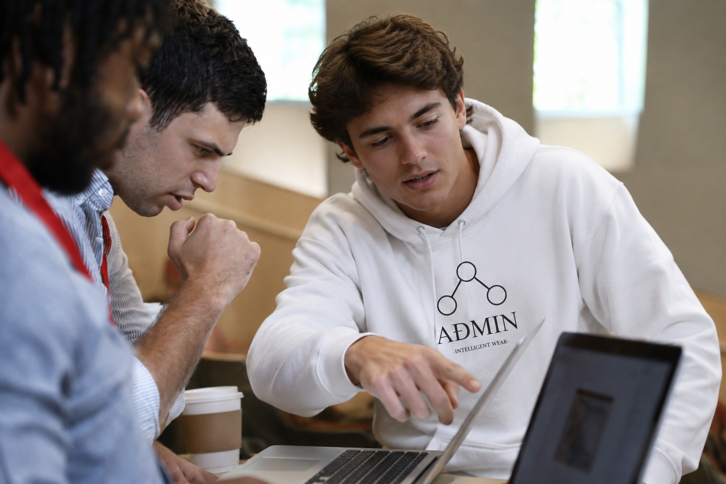A young man in a white hoodie with 'ADMIN' printed on the chest points at a laptop screen while sitting with two other men in a casual indoor setting.